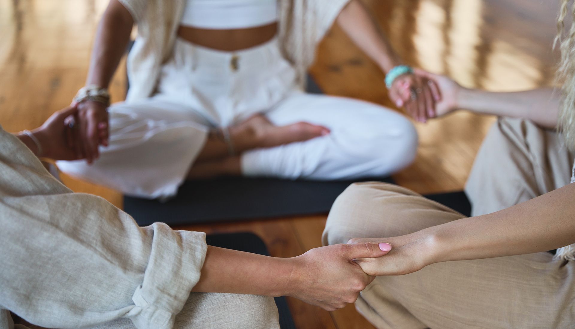 People in a circle meditating, holding hands. One person is in lotus position, wearing white. Others wear tan.