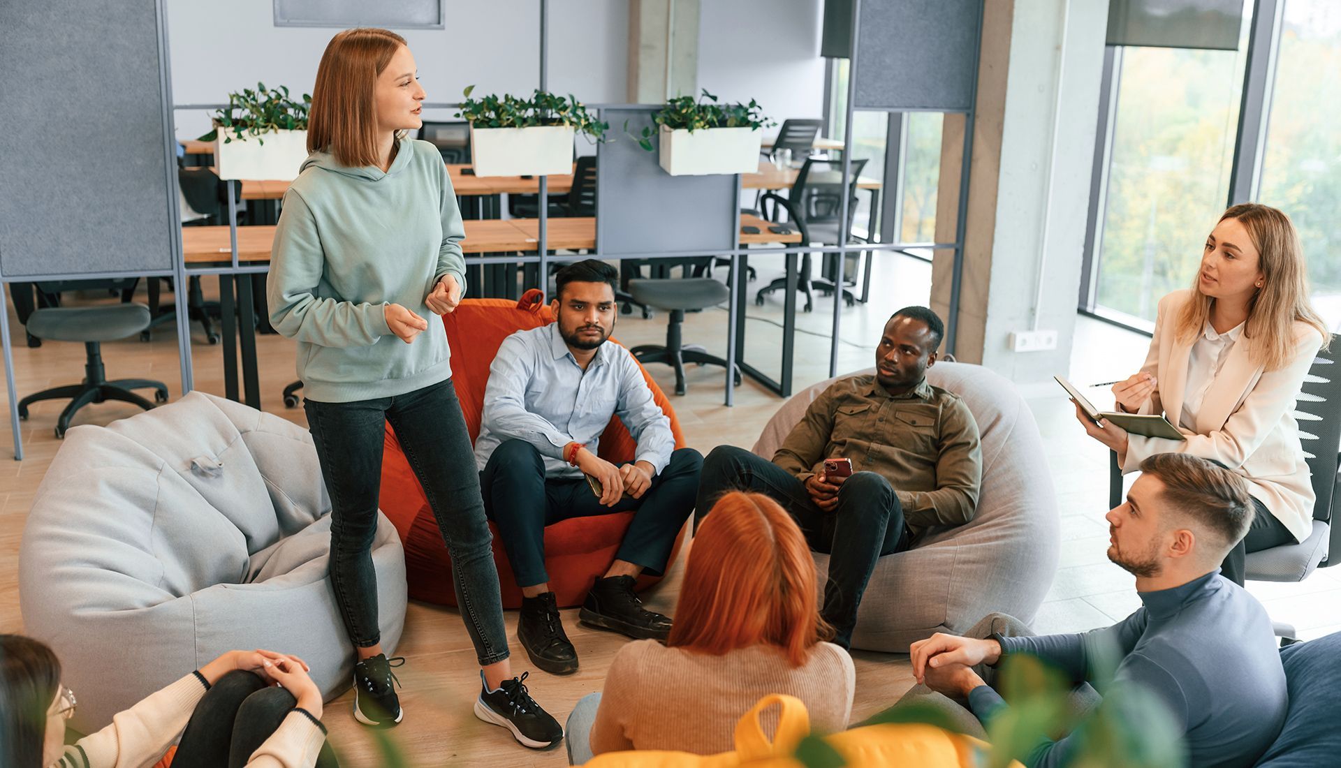 Group of people in casual setting; one standing, speaking; others seated on beanbags and chairs, listening.