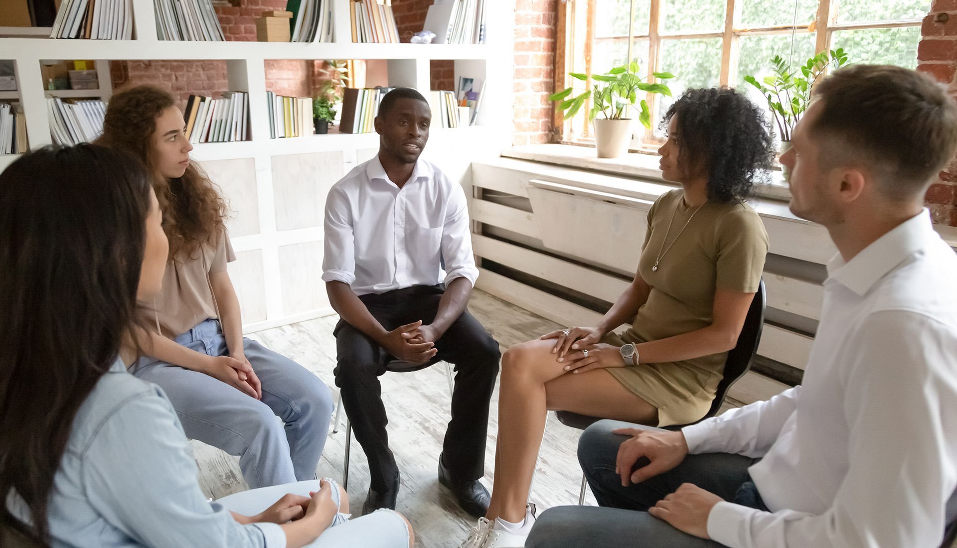 People in a circle seated, discussing. The central man in a white shirt is speaking. Natural light fills the room.