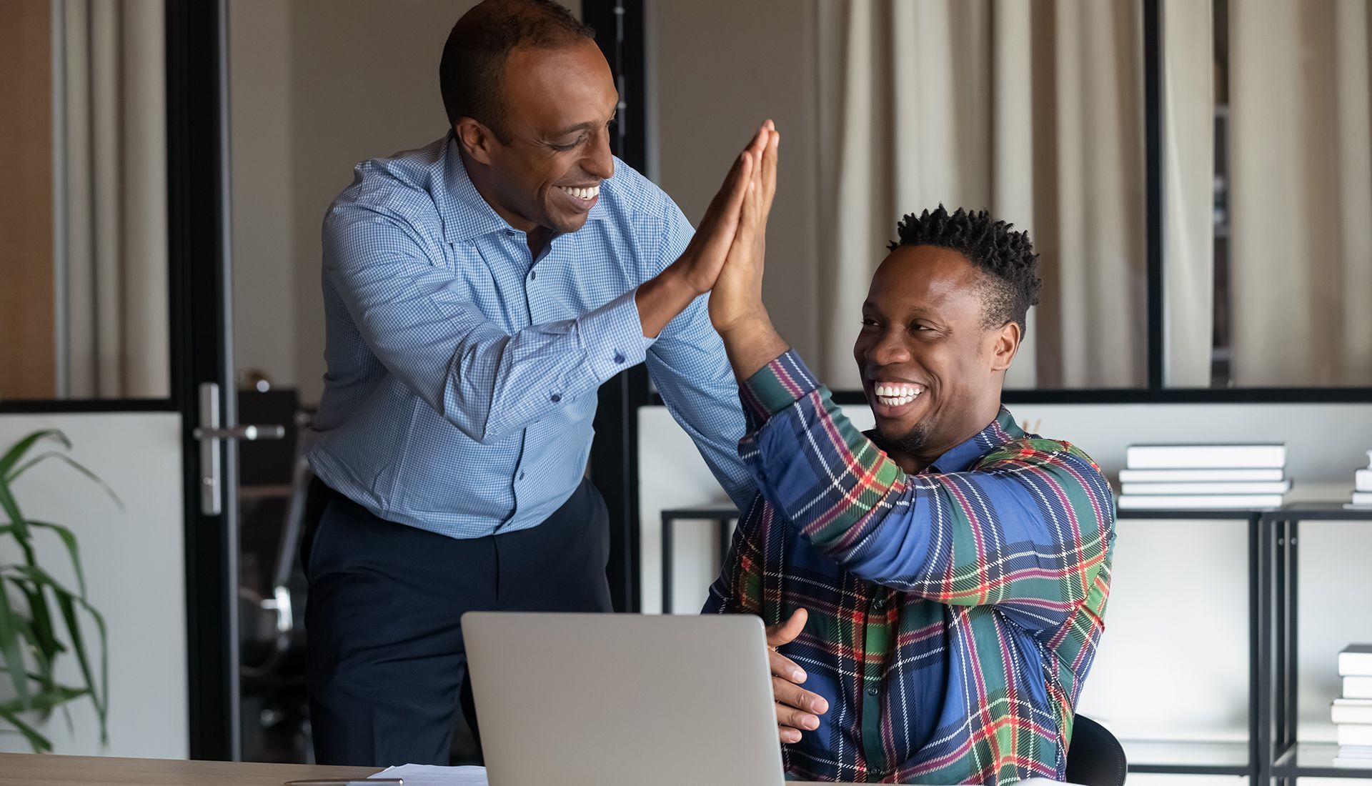 Two men high-fiving in office, smiling. One is seated at a laptop.