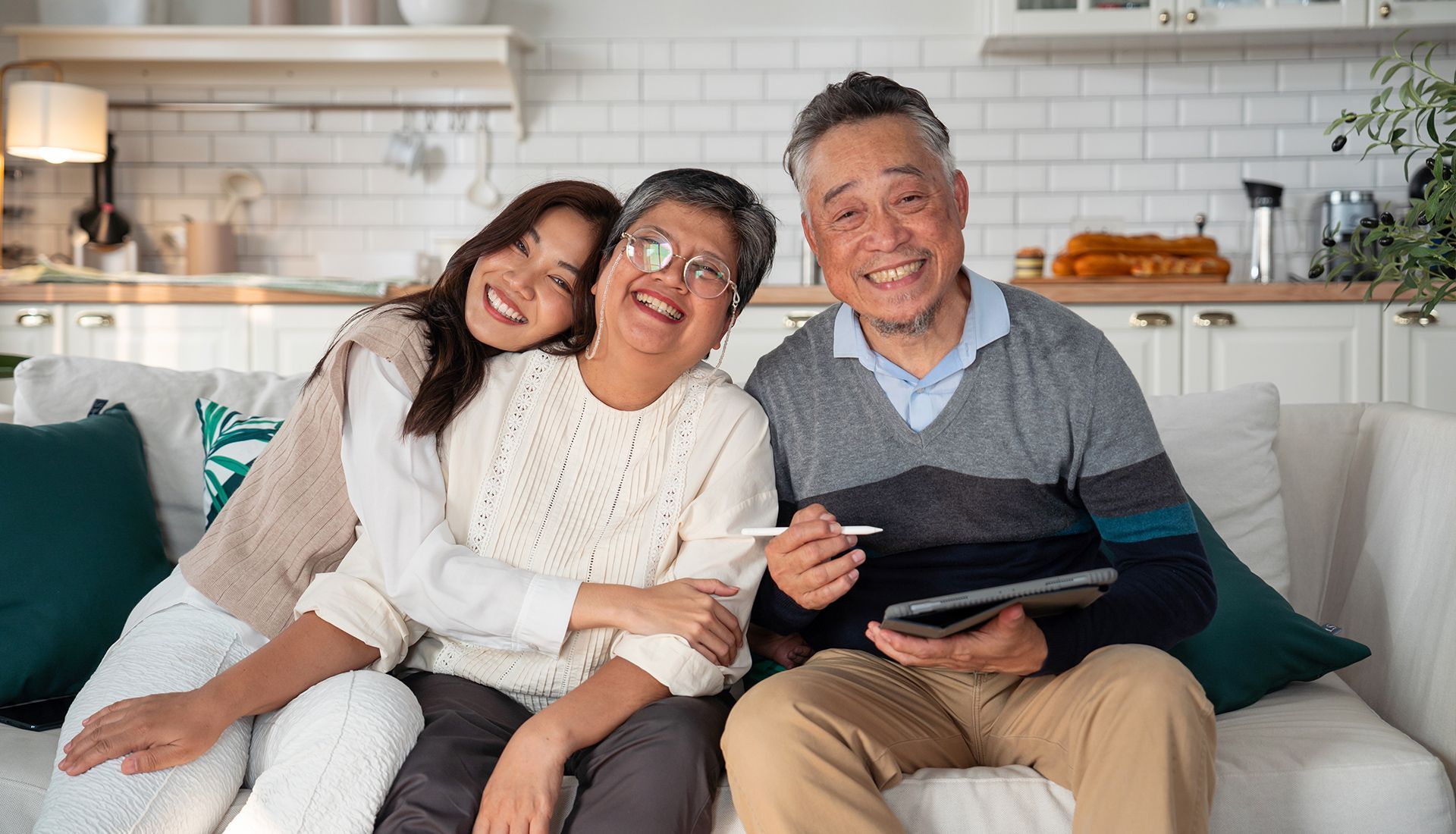 Woman hugs smiling older couple on a sofa, one holds a tablet in a bright kitchen.