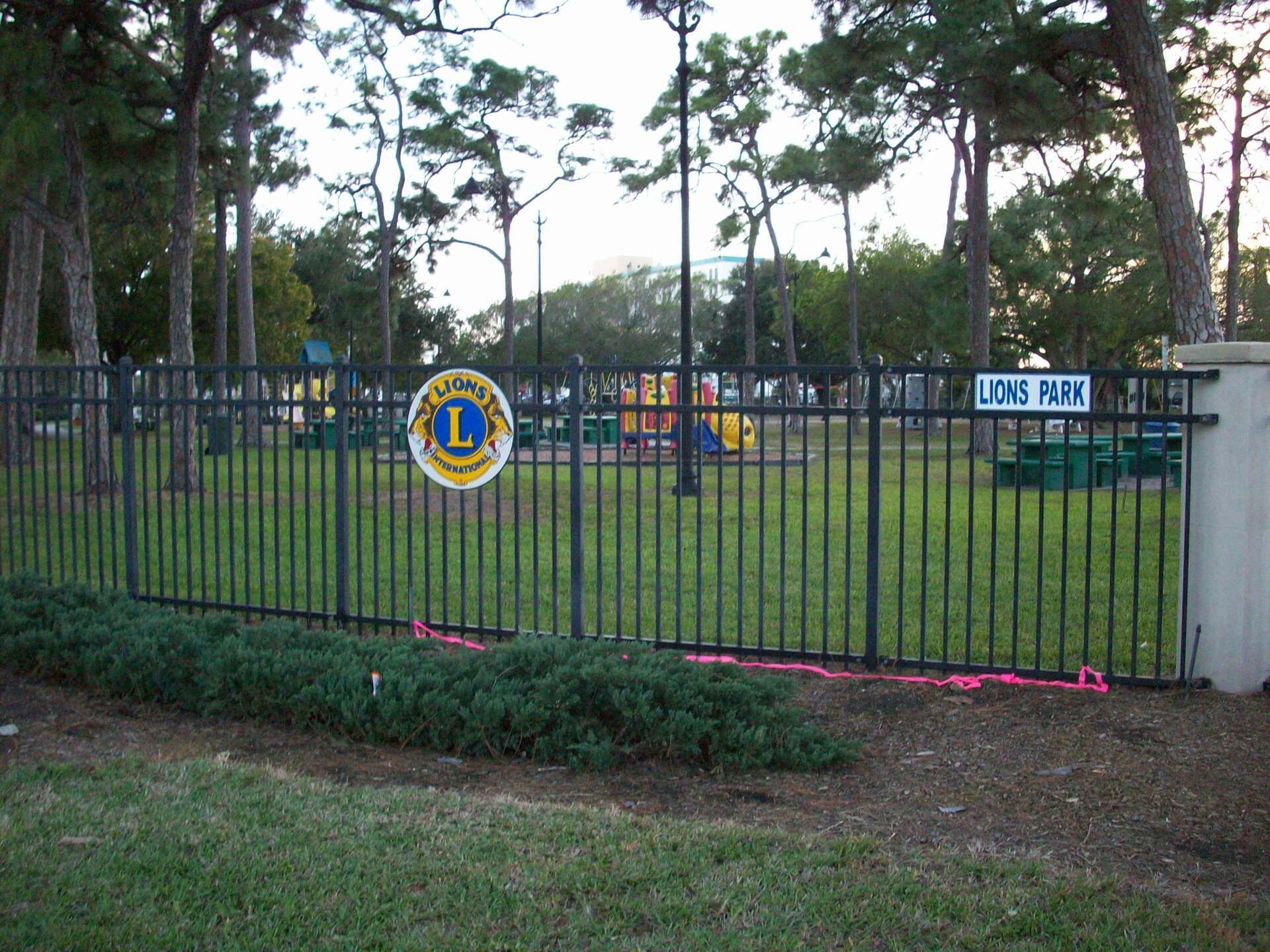 Black fence encloses Lions Park playground; trees in background. Sign with Lions Club logo.