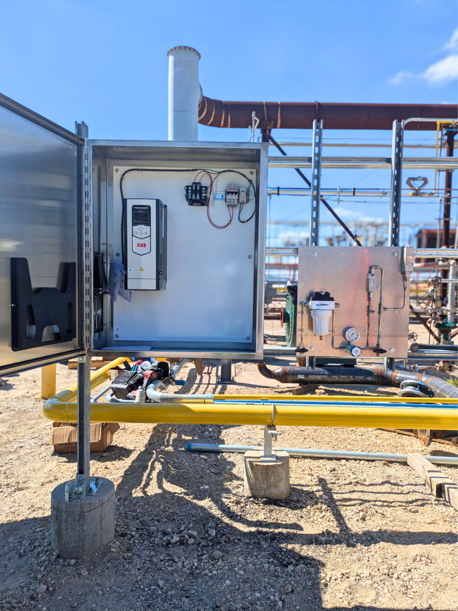 Open electrical panel with controls, mounted outdoors. Yellow pipes, metal framework, and concrete bases. Sunny day.