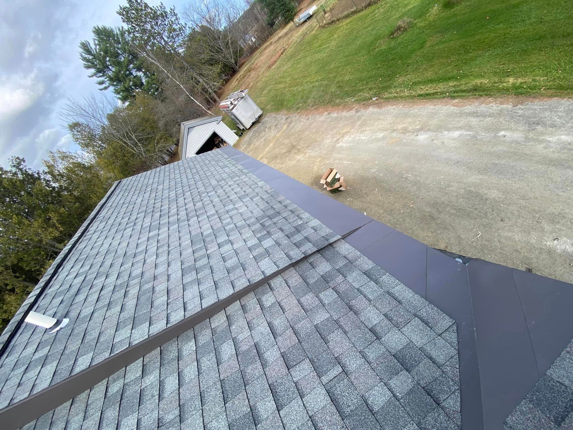 Gray shingled roof with a dark metal edge. Gravel driveway and grassy hill in the background. Overcast sky.