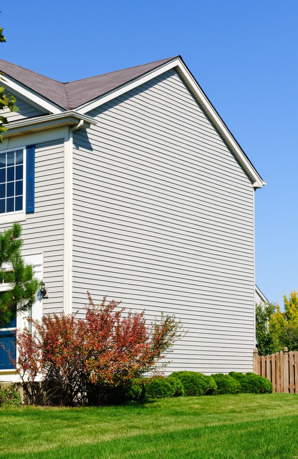 Two-story house with gray siding, blue shutters, and a red bush against a blue sky.