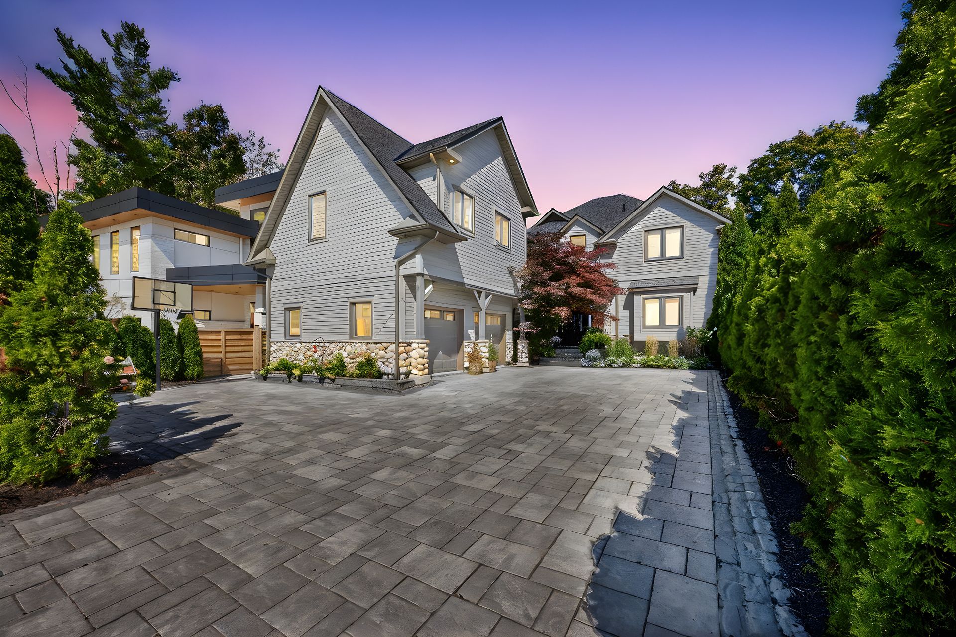 Modern gray and white house with brick driveway, surrounded by greenery at dusk.