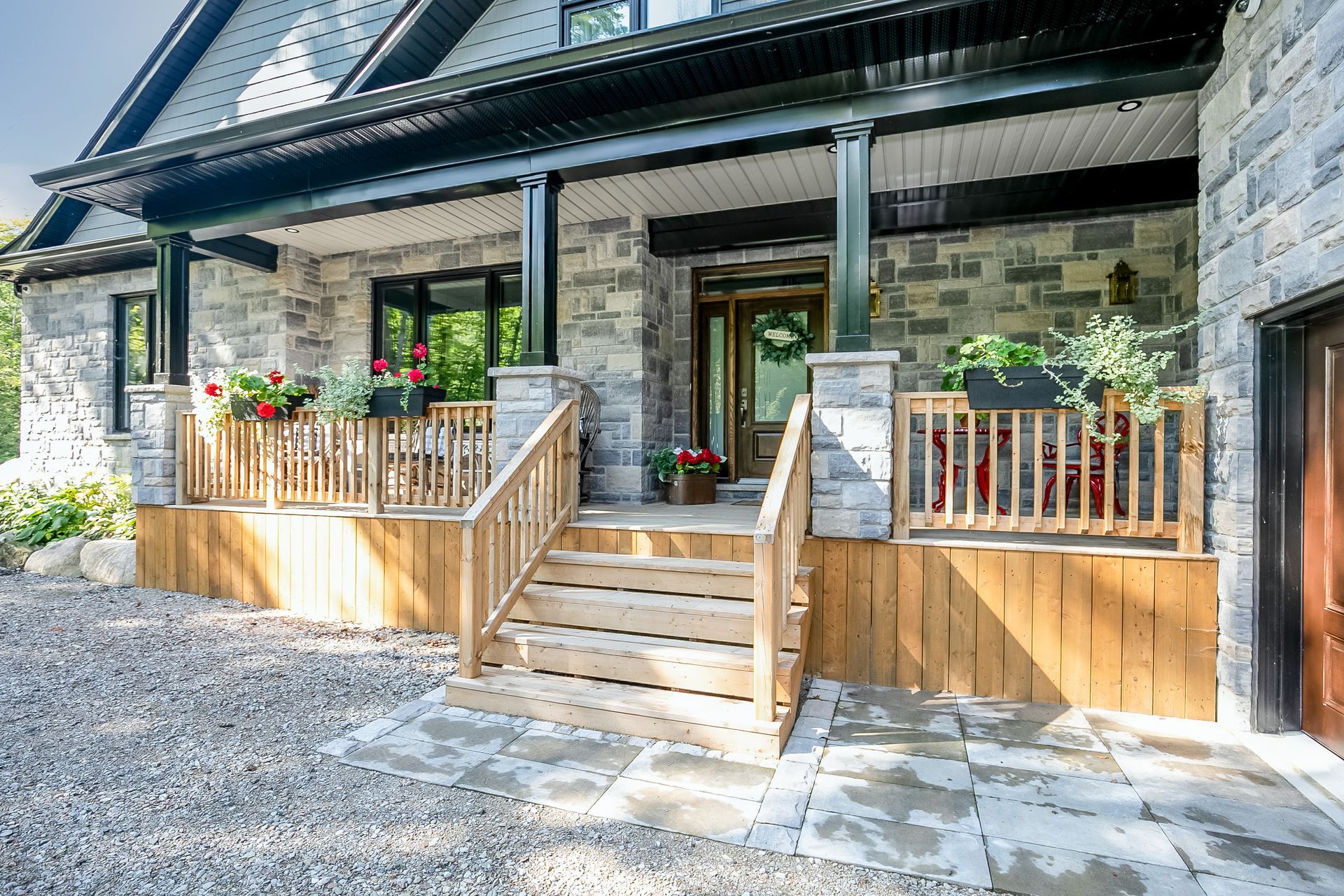 Stone house with wooden porch, steps, and railing. Red flowers and green wreath adorn the entrance.