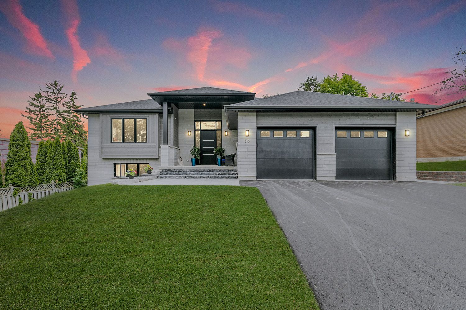 Modern gray house with black garage doors, driveway, and lawn under a pink and orange sunset.