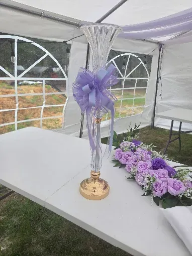 Purple ribboned centerpiece on a white table inside a tent, with lilac flowers beside it.