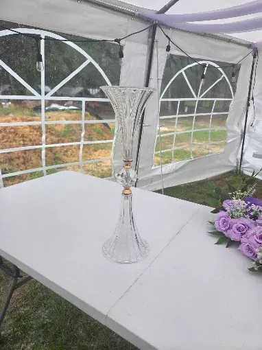 Clear glass vase on a white table inside a tent, with purple flowers nearby and windowed walls in back.