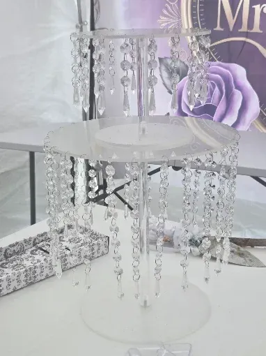 White acrylic cake stand with hanging crystal beads on a white tabletop beside a purple floral backdrop