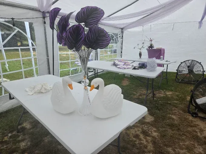 White folding tables in a decorated tent with purple floral centerpieces and swan napkin sculptures