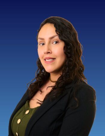 A person with long dark wavy hair, wearing an olive green button-up top and black blazer against a blue gradient background.