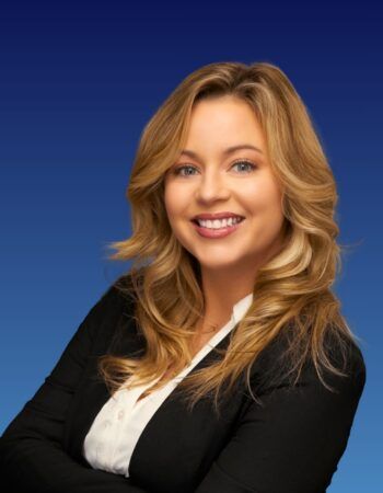 A professional headshot of a smiling woman with wavy blonde hair, wearing a white shirt and black blazer against a blue.
