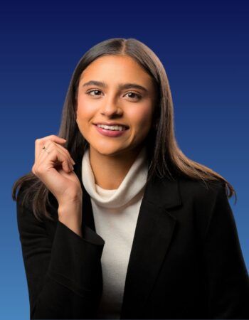 A person with long dark hair wearing a white turtleneck and black blazer, smiling against a solid blue background.