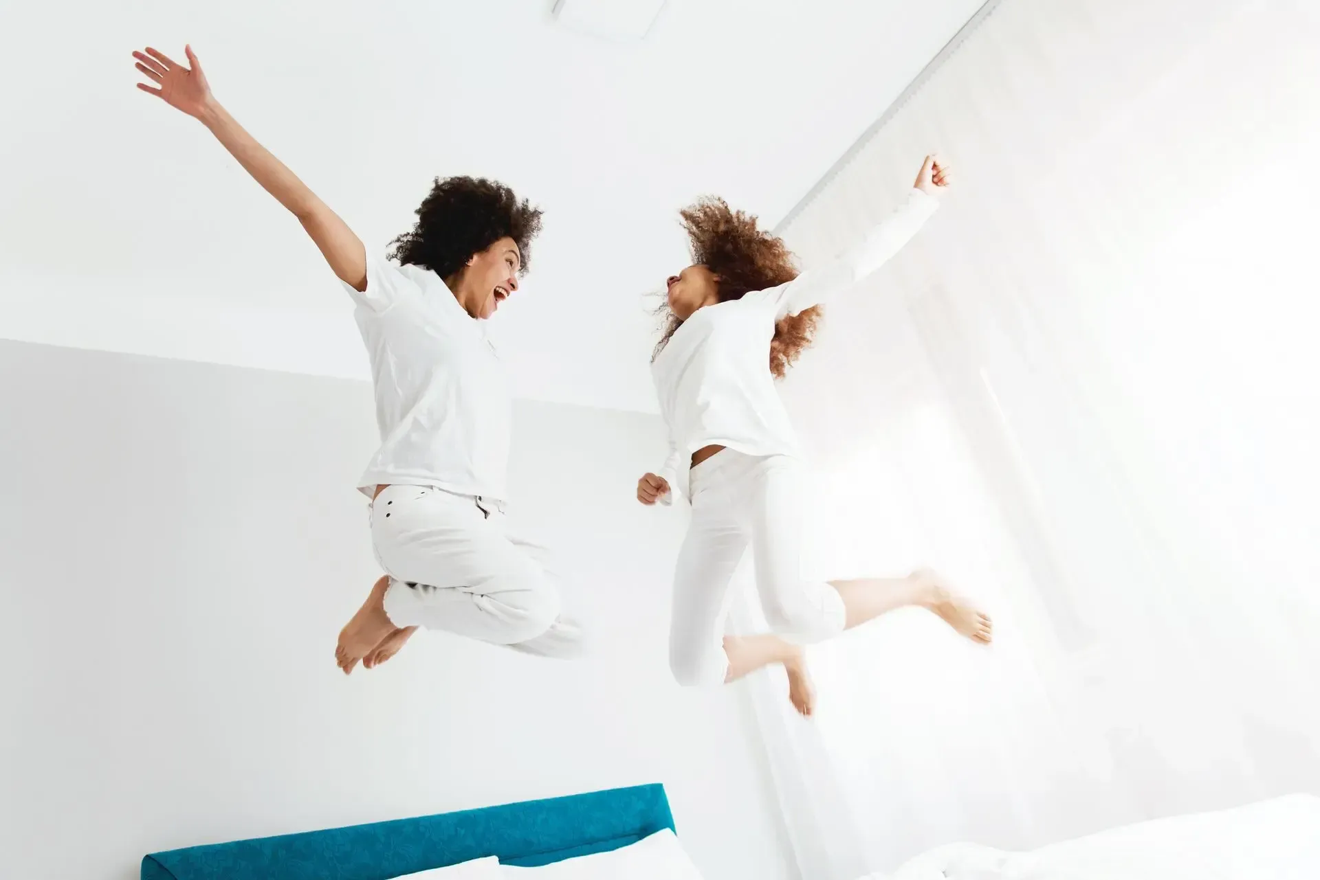 Two people in white clothes jump joyfully in a bright white room near a blue couch.