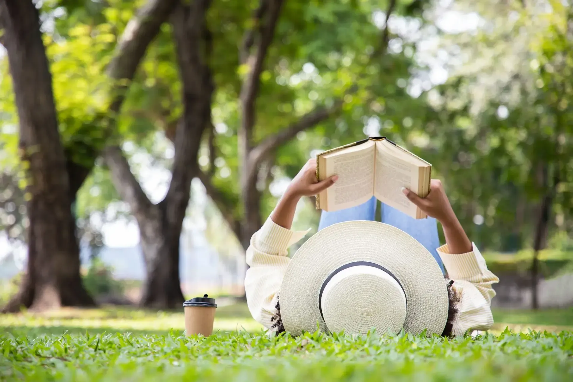 Person lying on grass reading a book under a sunhat in a park, with a coffee cup nearby.