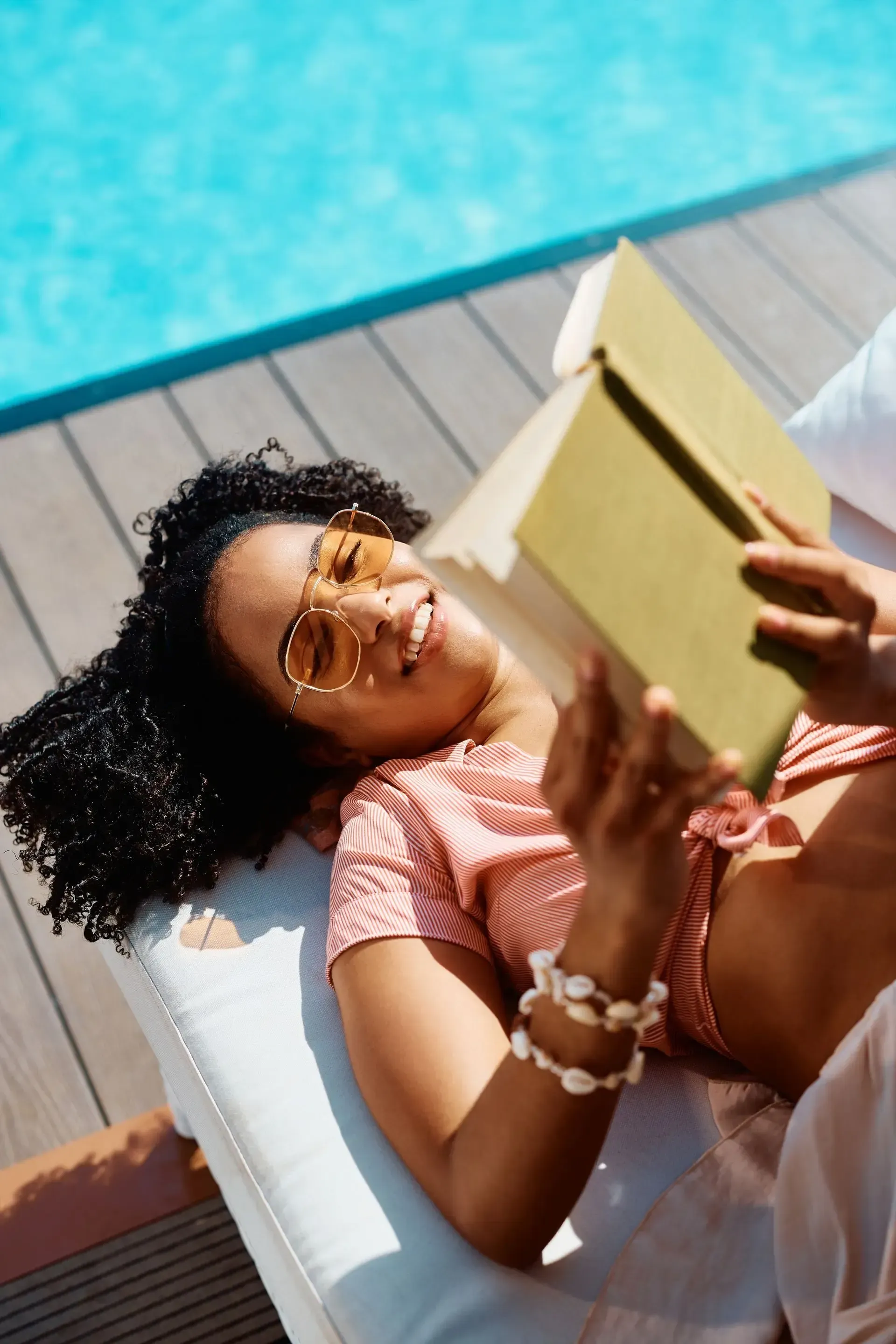 Person relaxing on a lounge chair by a pool, reading a book in bright sunlight.