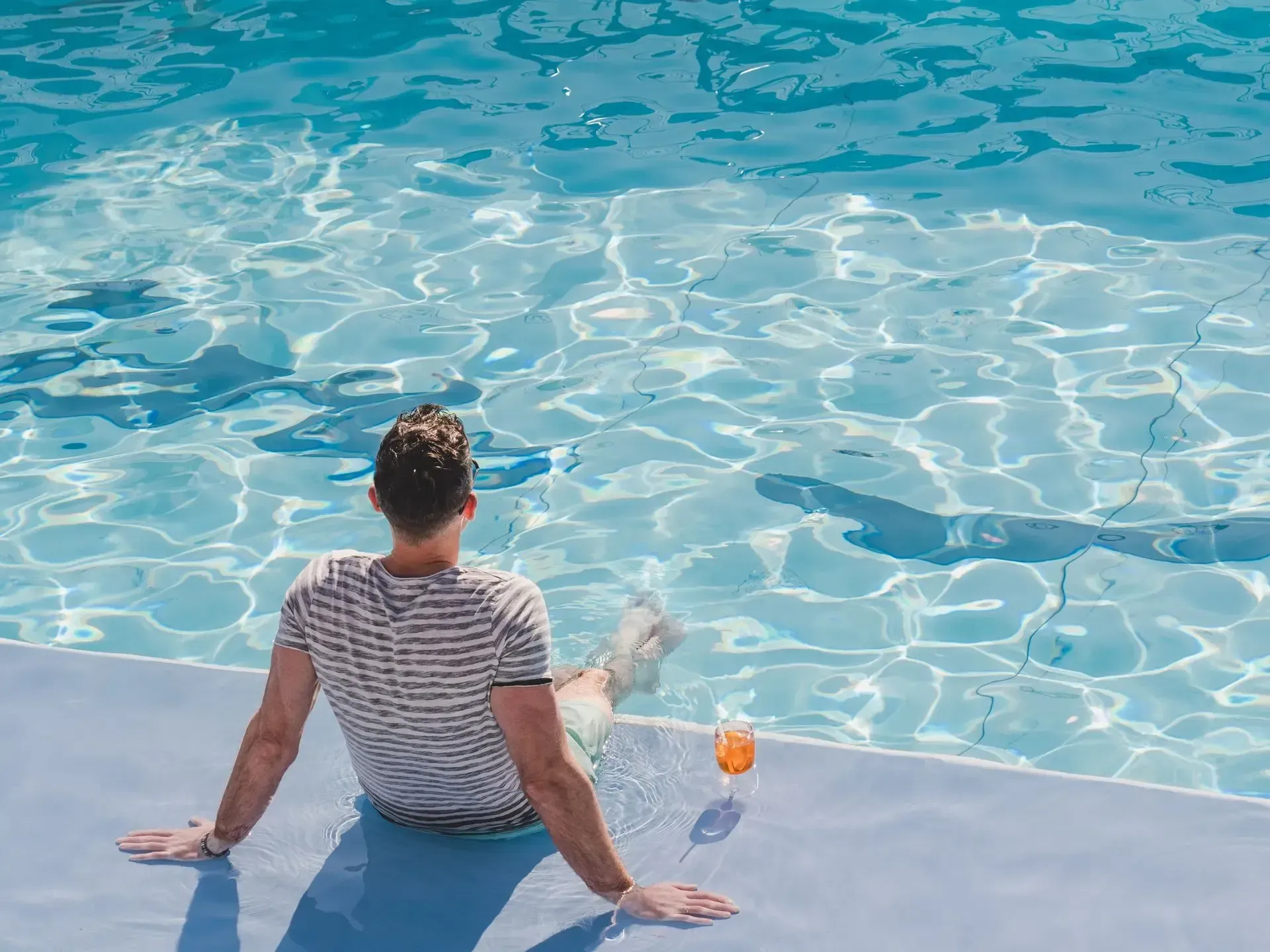 Person sitting at the edge of a pool, facing the water with a drink beside them