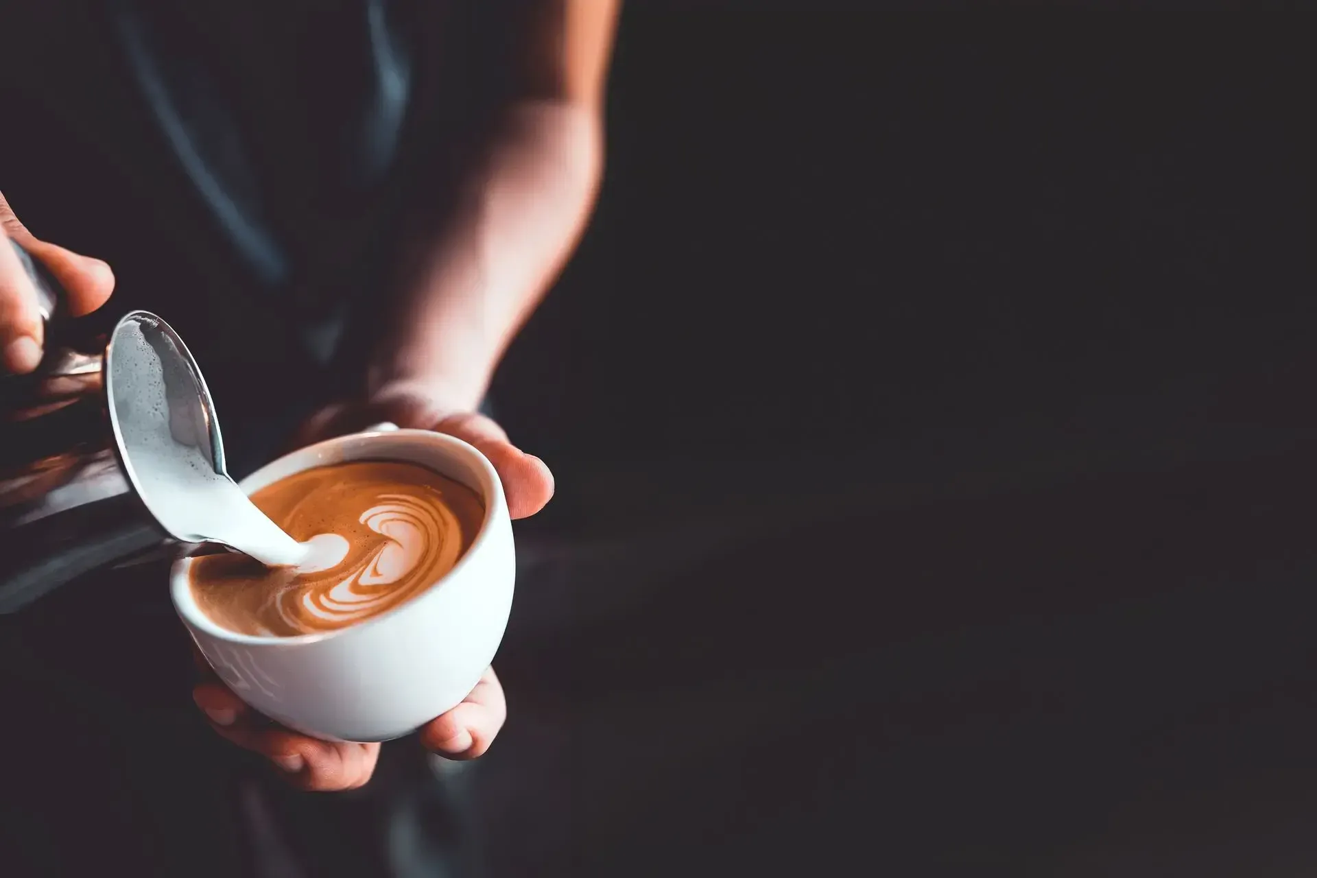 Hands holding a white cup of coffee with latte art and a spoon over a dark background