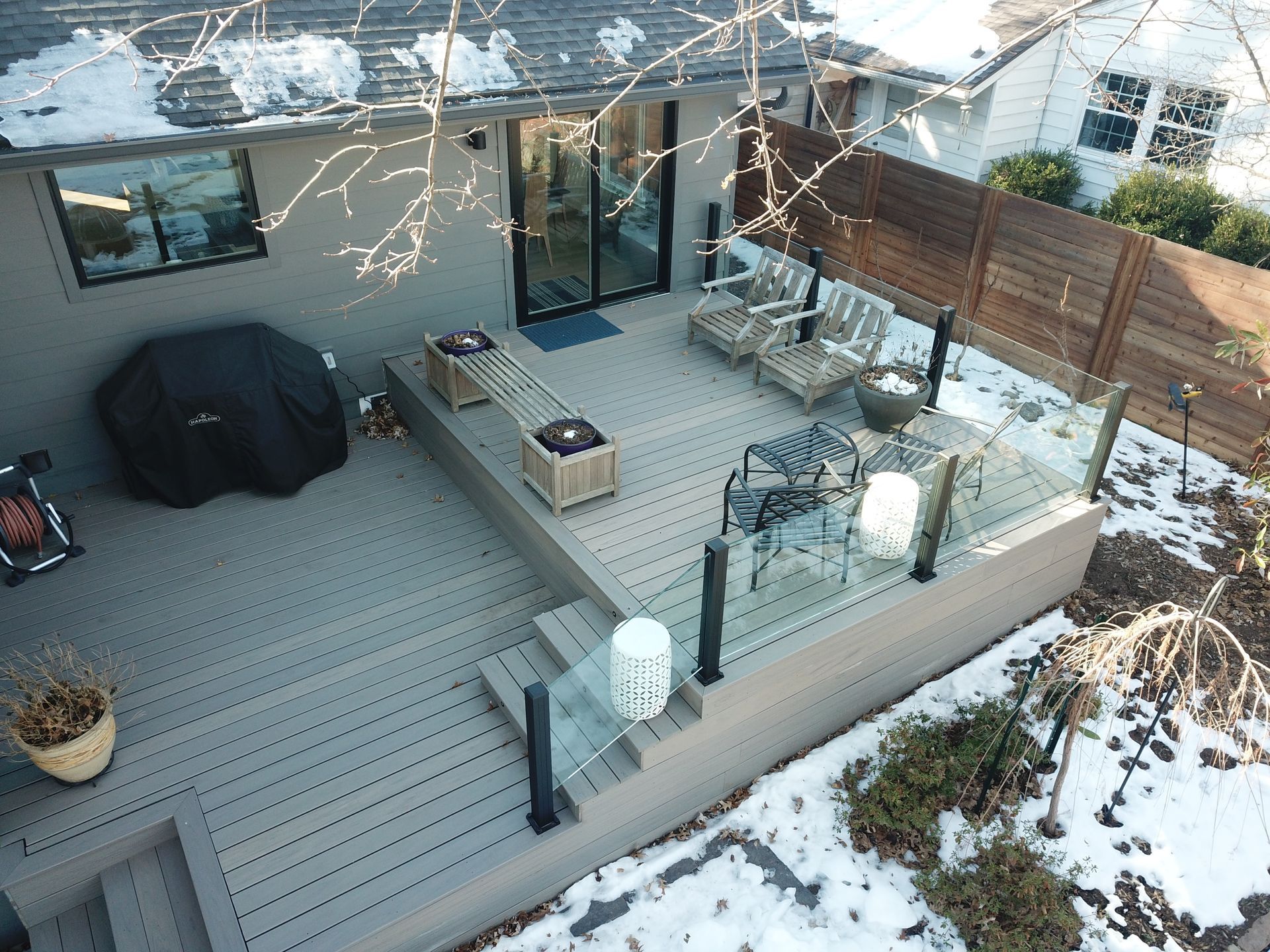 Backyard deck with outdoor seating, grill, glass railing, and patches of snow surrounding it