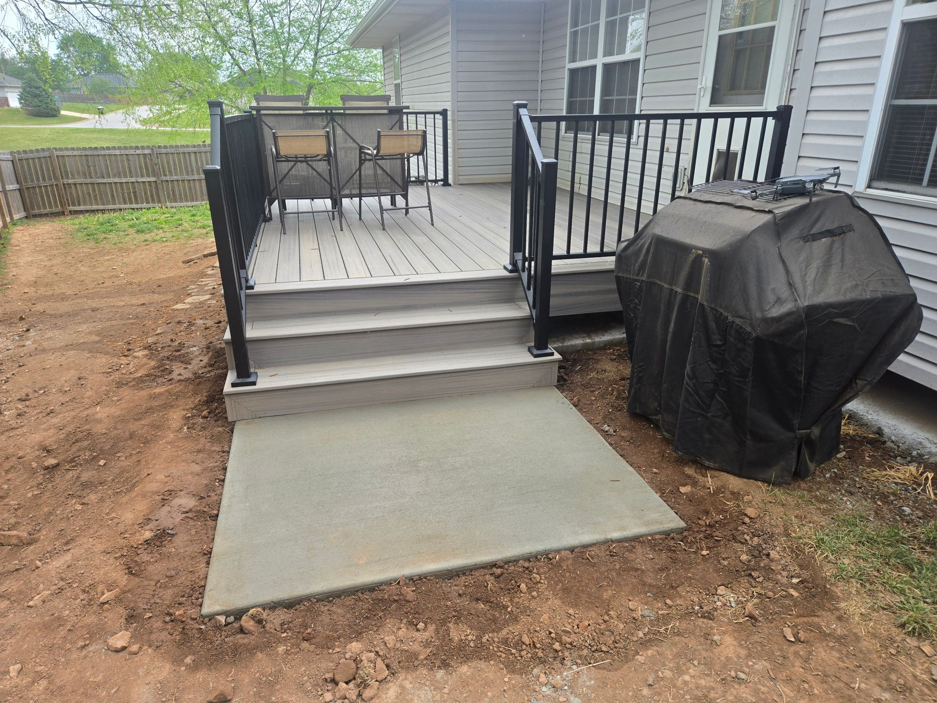 Backyard deck with steps, black railings, patio furniture, and a covered grill beside a concrete pad