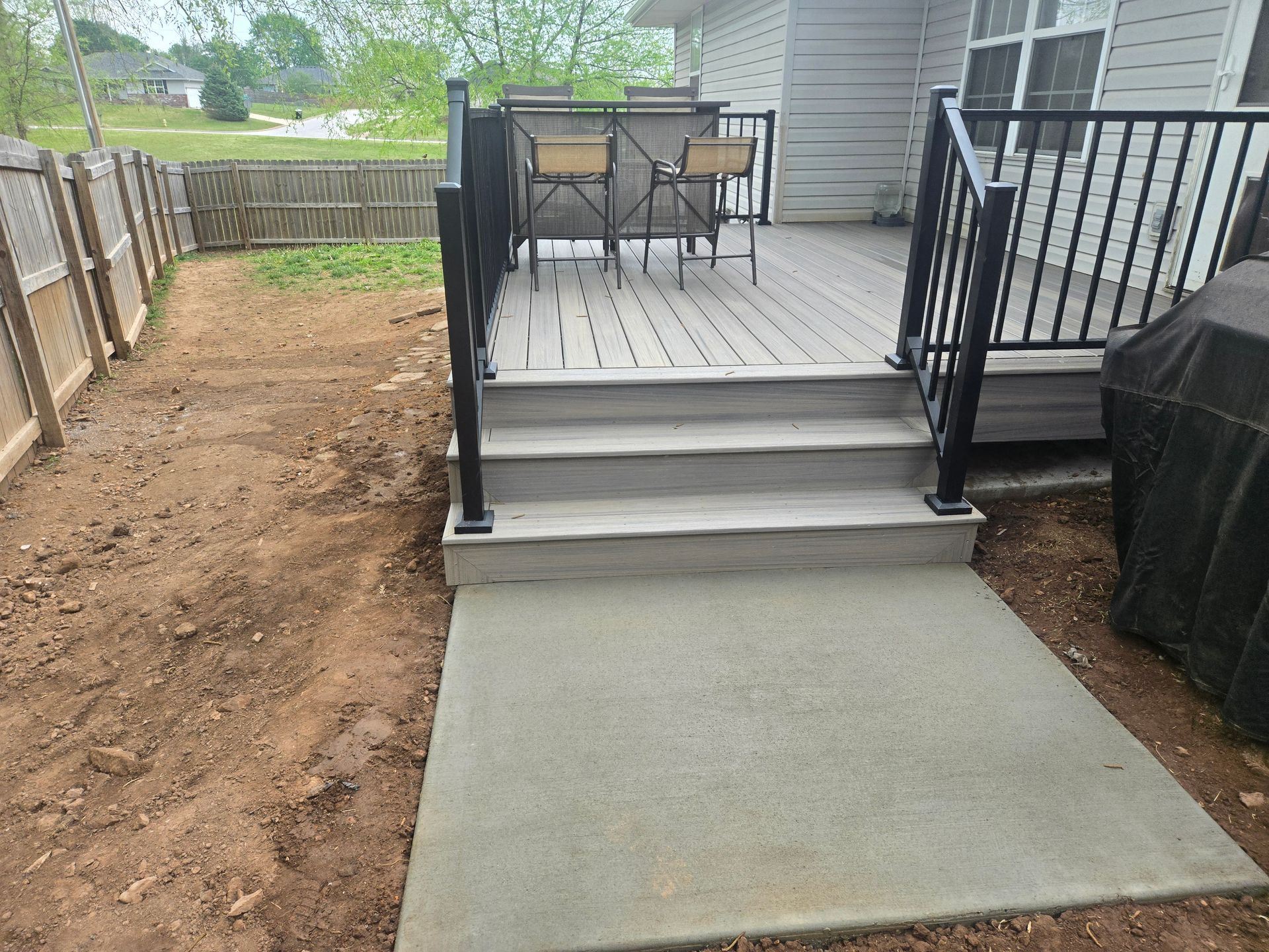 Concrete path to a backyard deck with black railings and outdoor seating, beside a fenced yard.