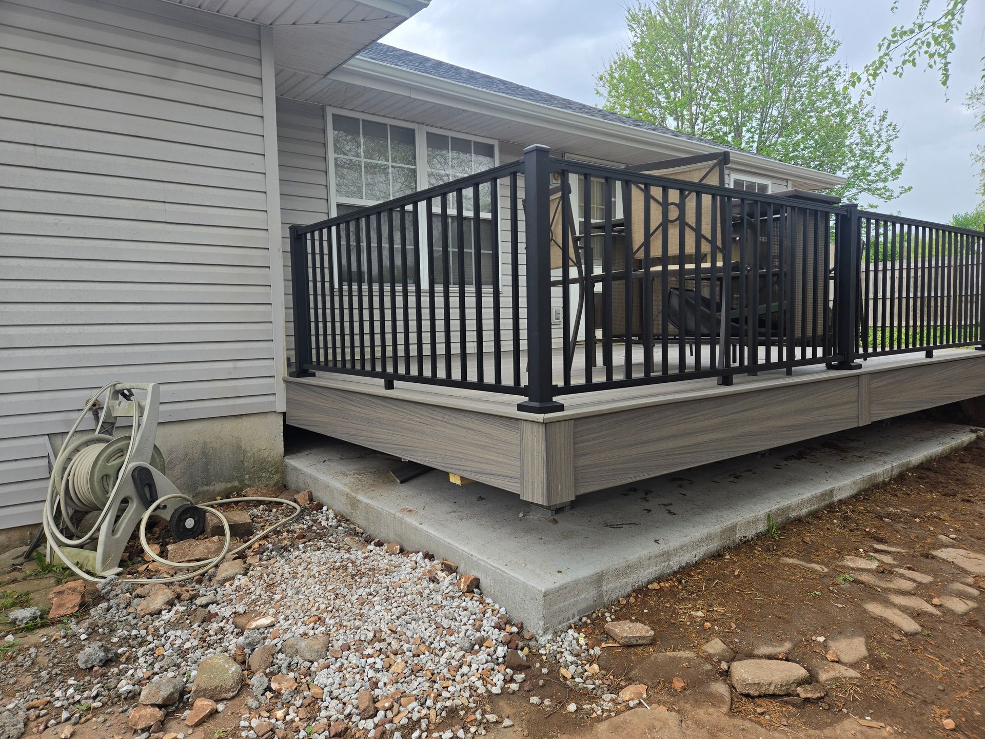 Raised wooden deck with black railing beside a house, on a concrete foundation in a gravel yard