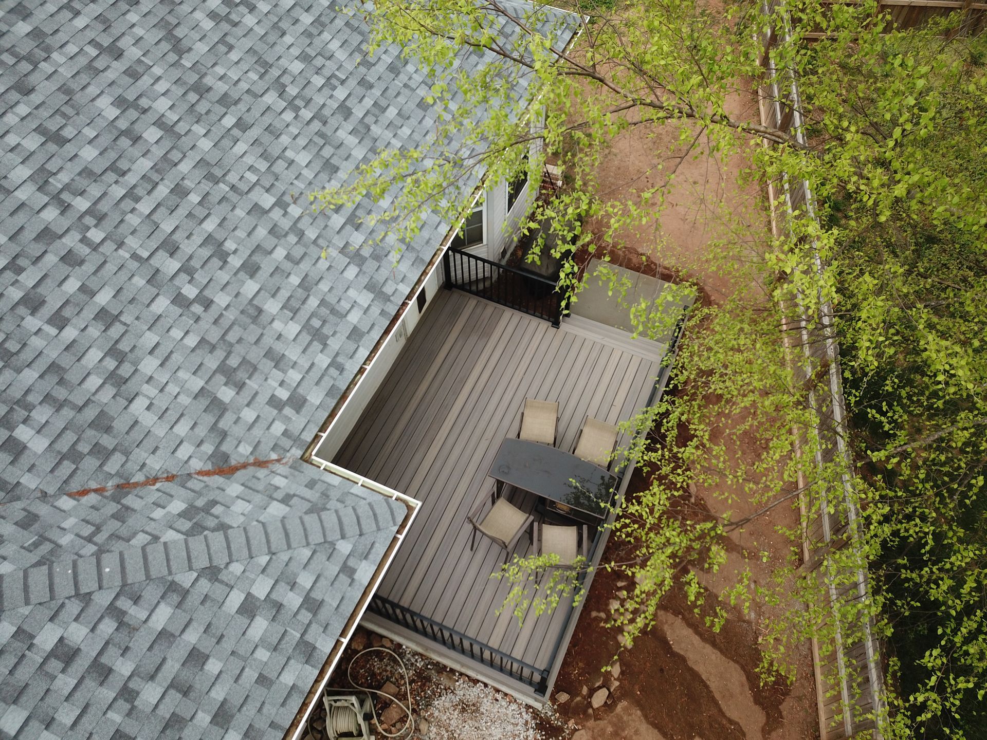 Overhead view of a rooftop deck with a table and chairs beside a gray shingled roof and green trees.