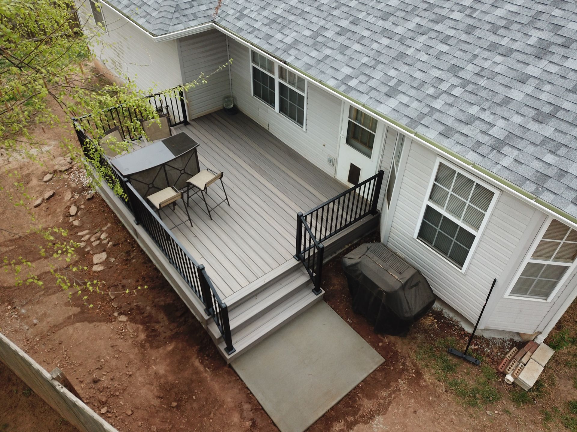 Back porch with gray deck, black railings, patio furniture, and a grill beside a white house.