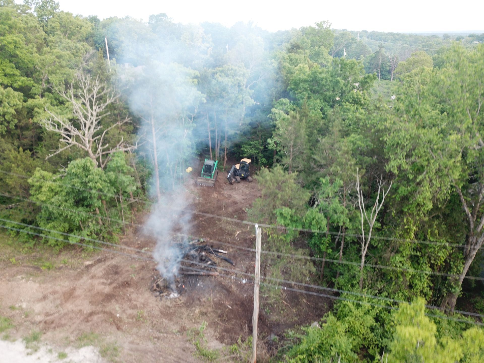 Smoke rising from a small ground fire at the edge of a wooded clearing near power lines.