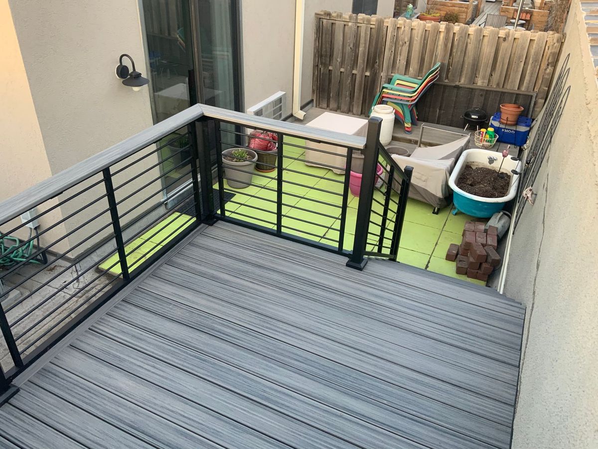 Small rooftop patio with gray decking, black railings, green mats, and potted plants beside a wooden fence.