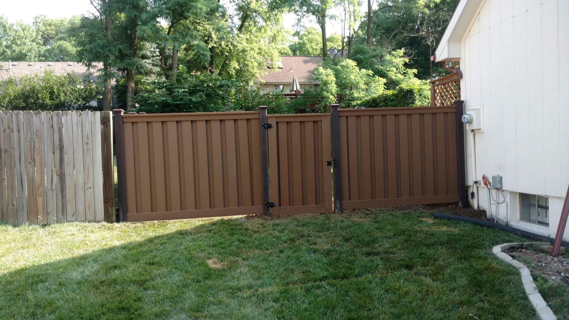 Backyard with a brown wooden fence, green grass, trees, and the side of a white house