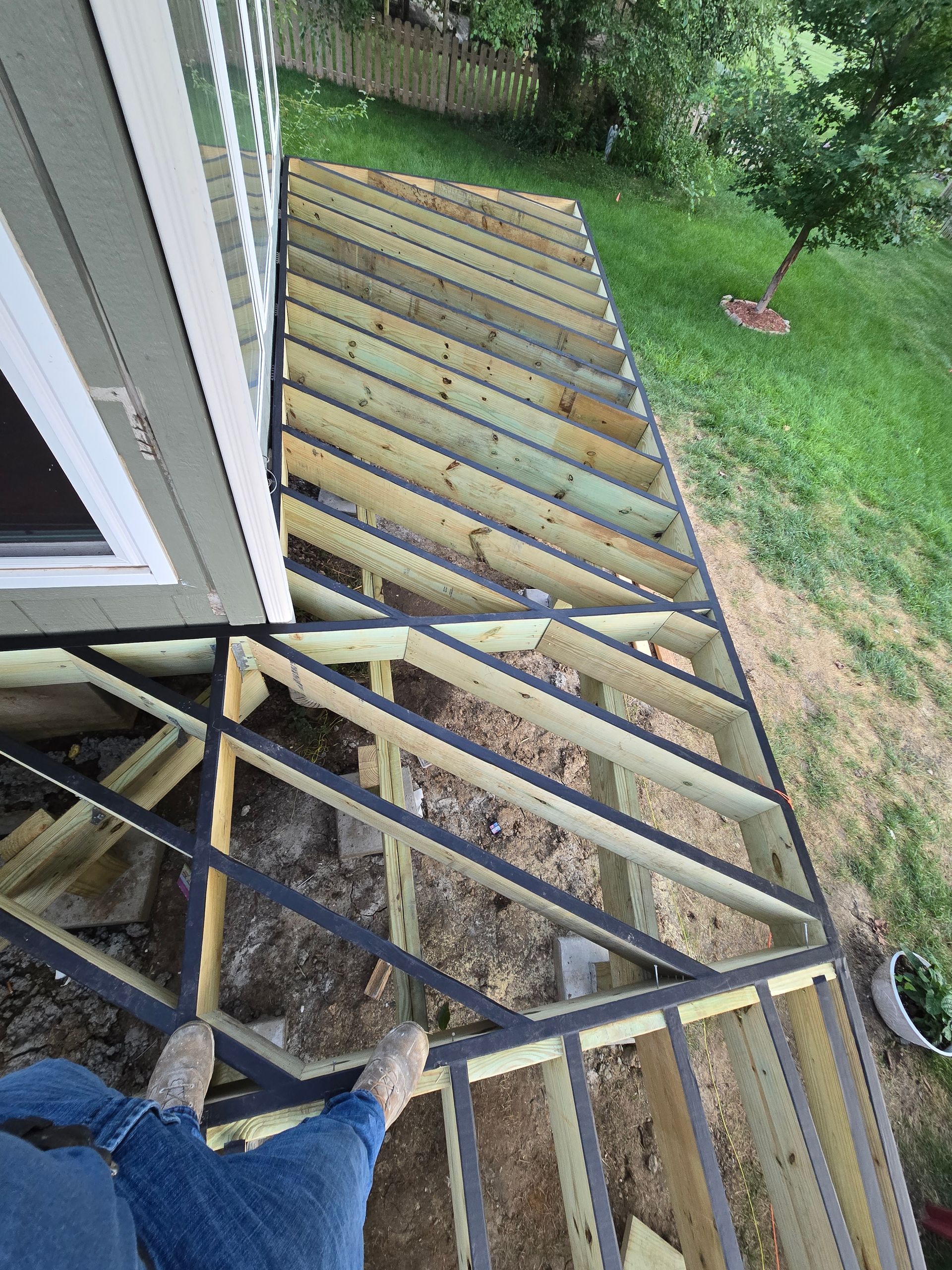 Worker on a roof deck framing, with wood joists beside a house and green lawn below.