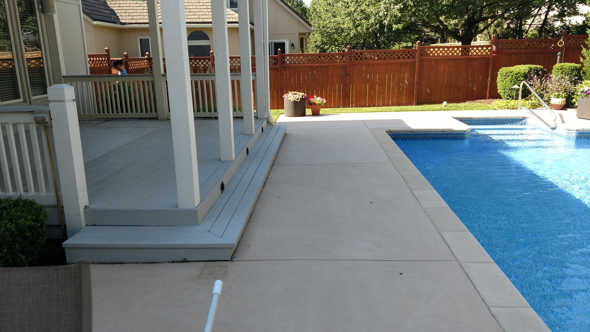 Poolside patio with white columns, concrete walkway, and blue swimming pool beside a wooden fence.