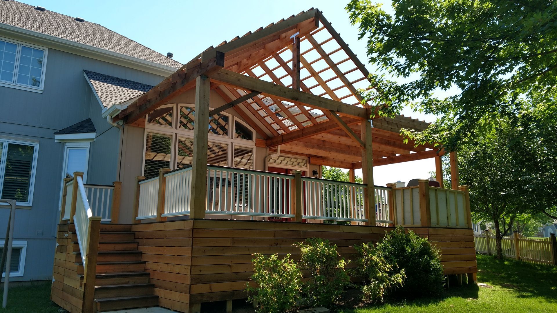 Wooden porch deck with a slatted roof attached to a blue house in a sunny backyard