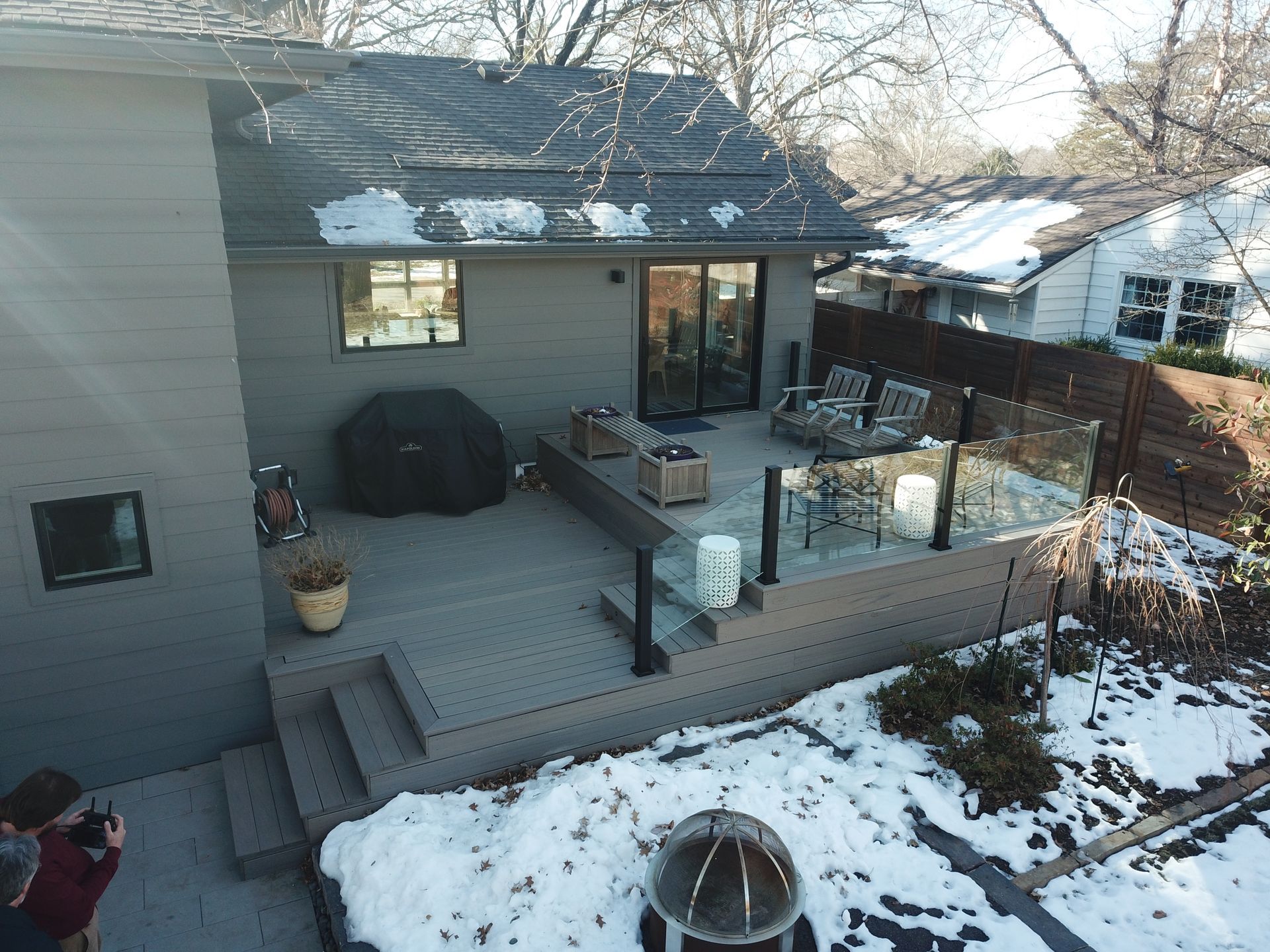 Snowy backyard deck with patio furniture, grill, and a fire pit beside a house