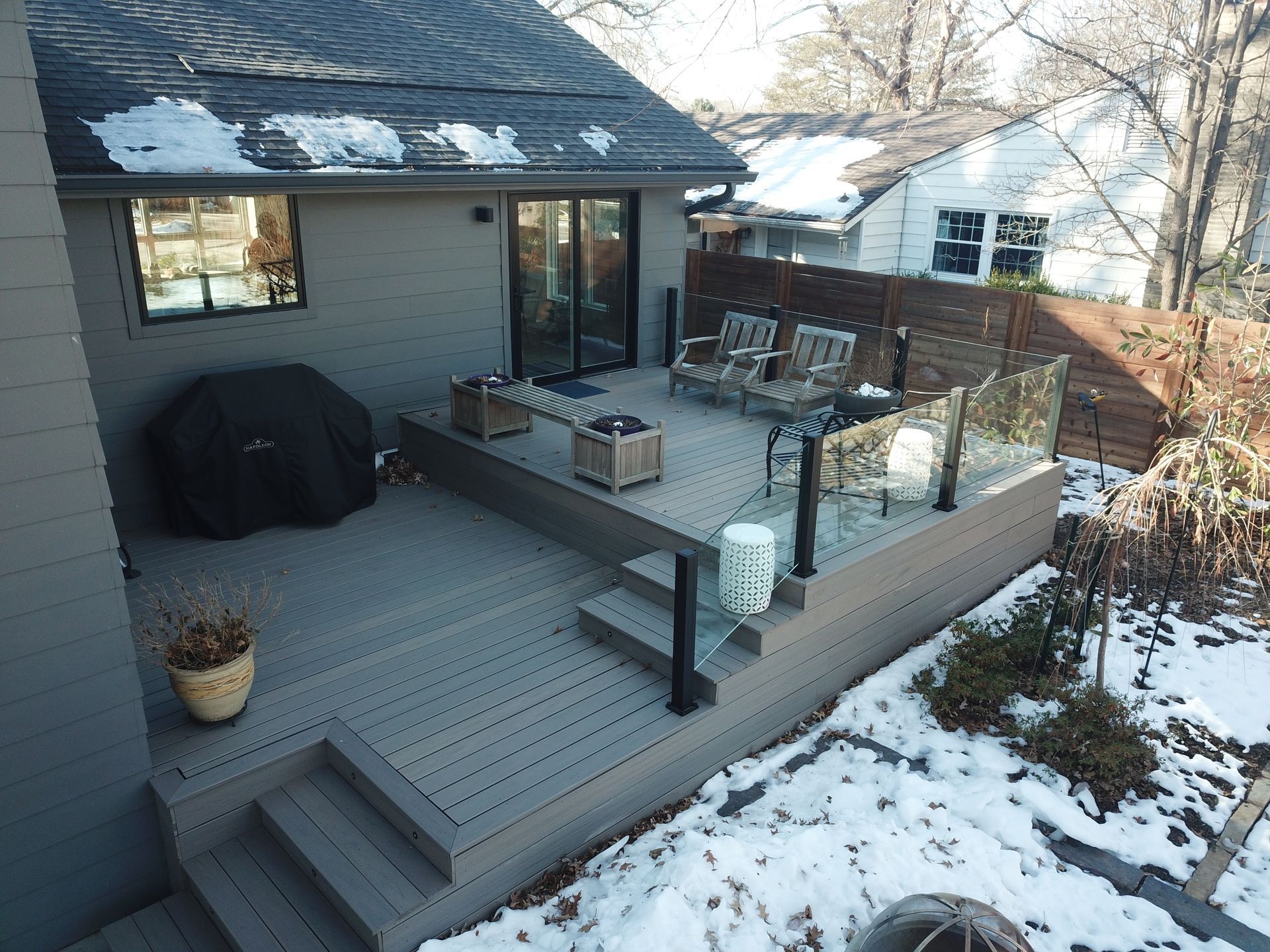 Snowy backyard deck with outdoor furniture beside a gray house and glass railing
