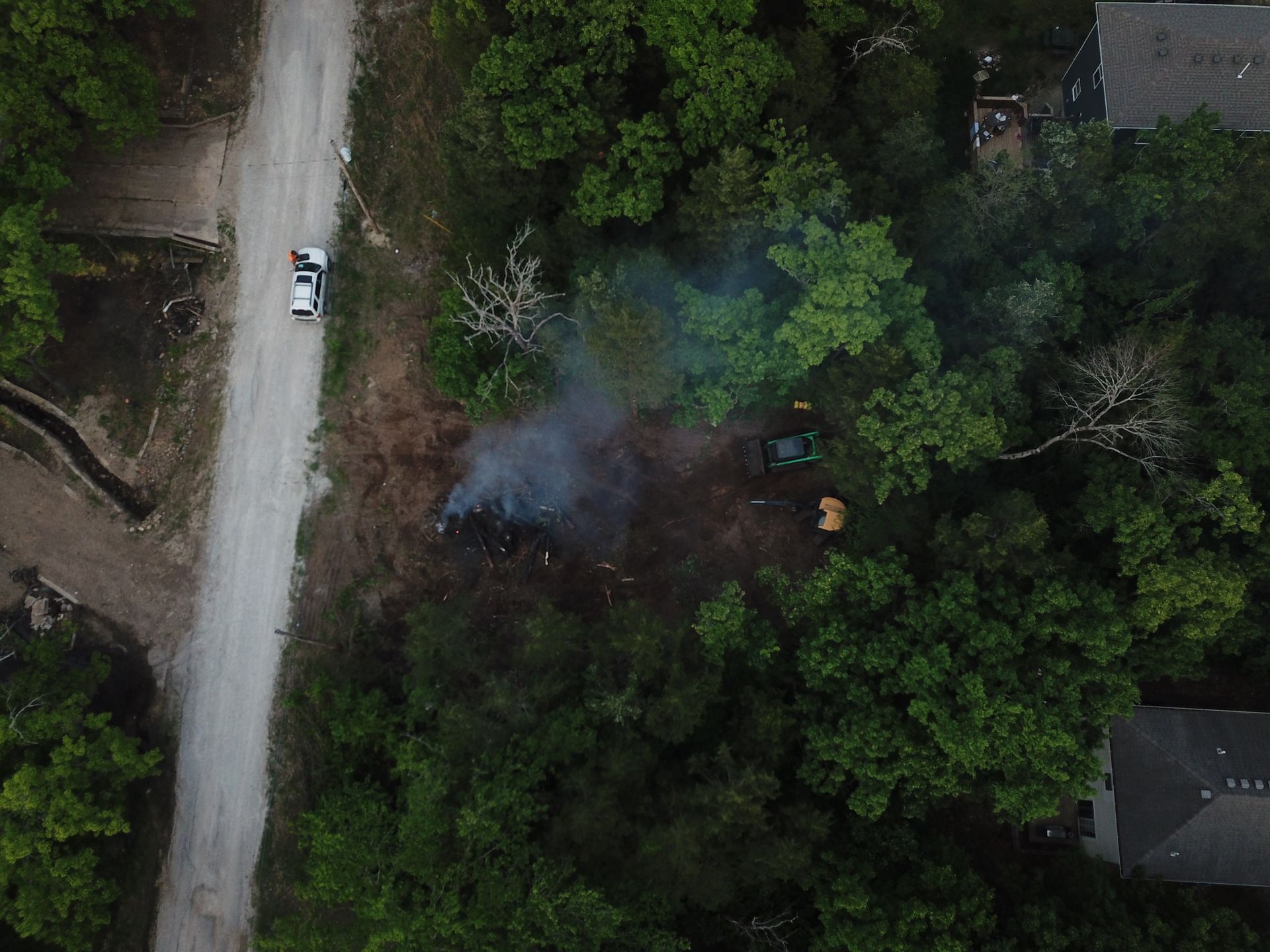 Aerial view of a smoky fire in dense green trees beside a dirt road and a parked vehicle.