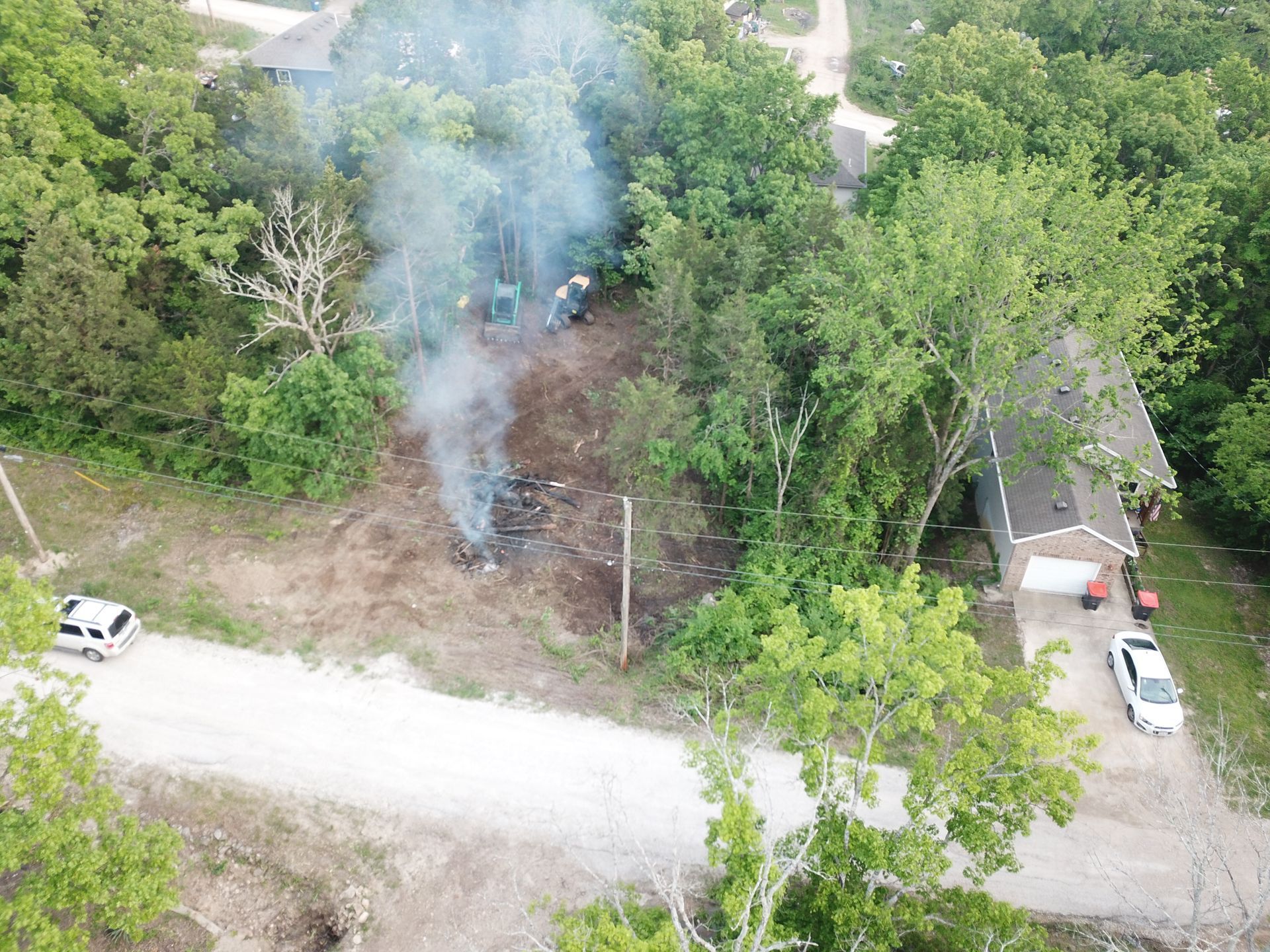 Aerial view of a smoky fire in a wooded clearing beside a road, with cars nearby.