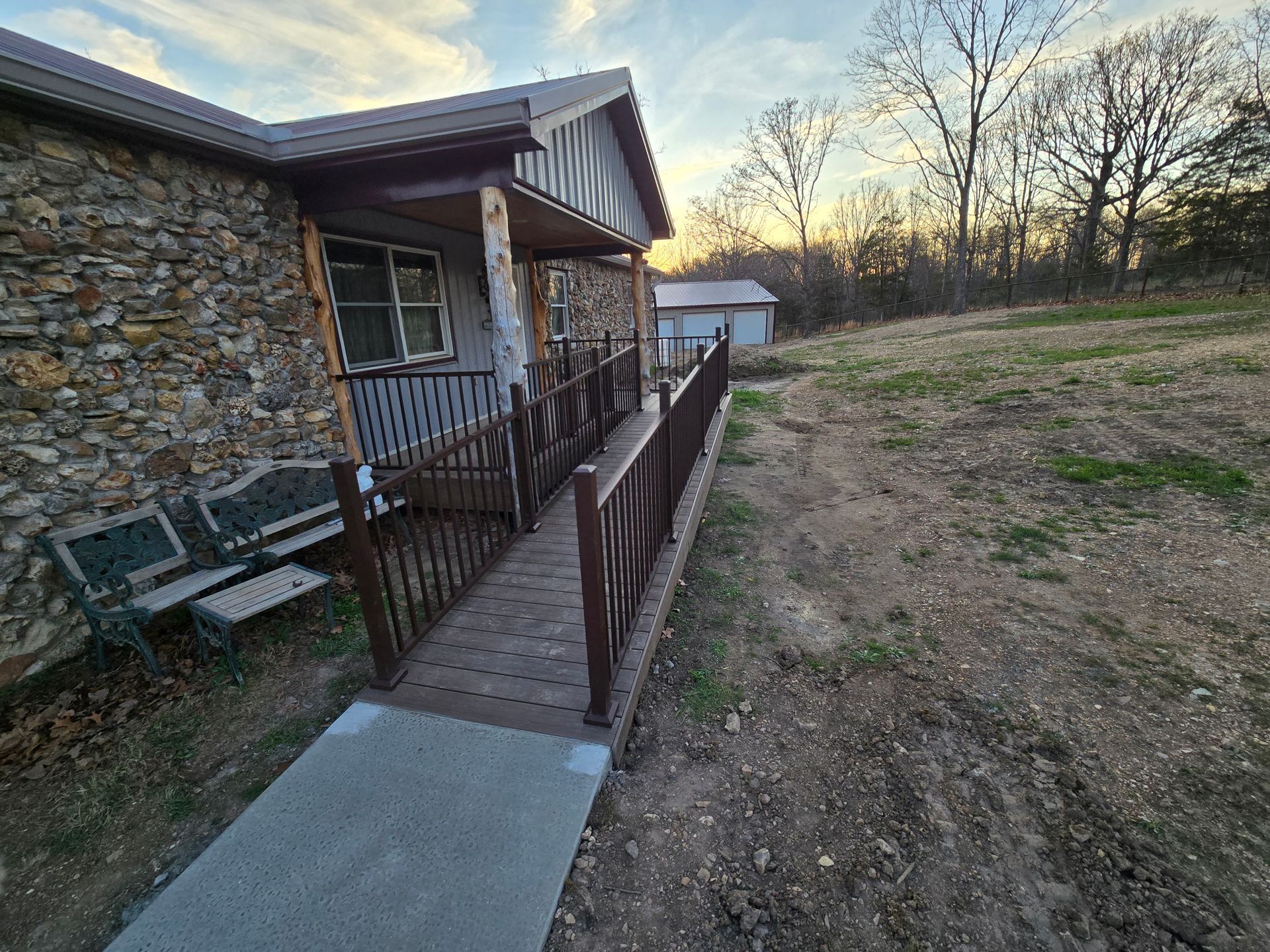 Ramped walkway beside a rustic stone house at sunset, leading to a grassy yard and small shed.