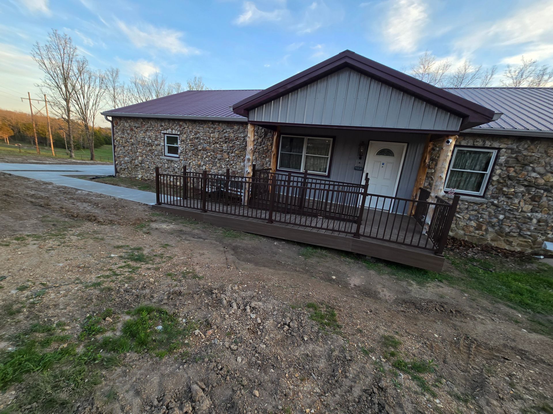 Stone building with a covered entrance and metal railing, beside a gravel driveway at sunset