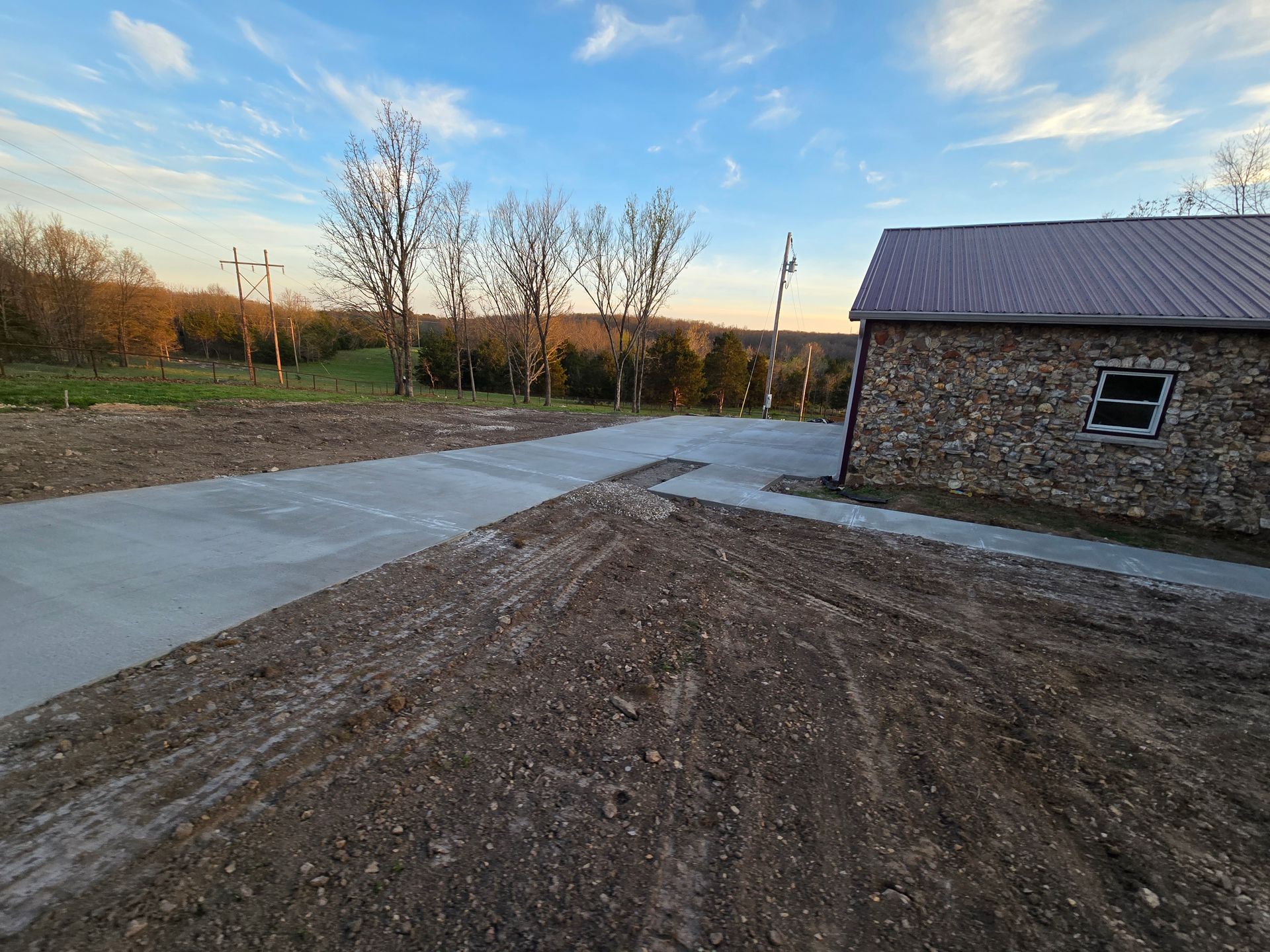 Dirt driveway beside a stone building at sunset, with bare trees and a concrete path leading into the distance.