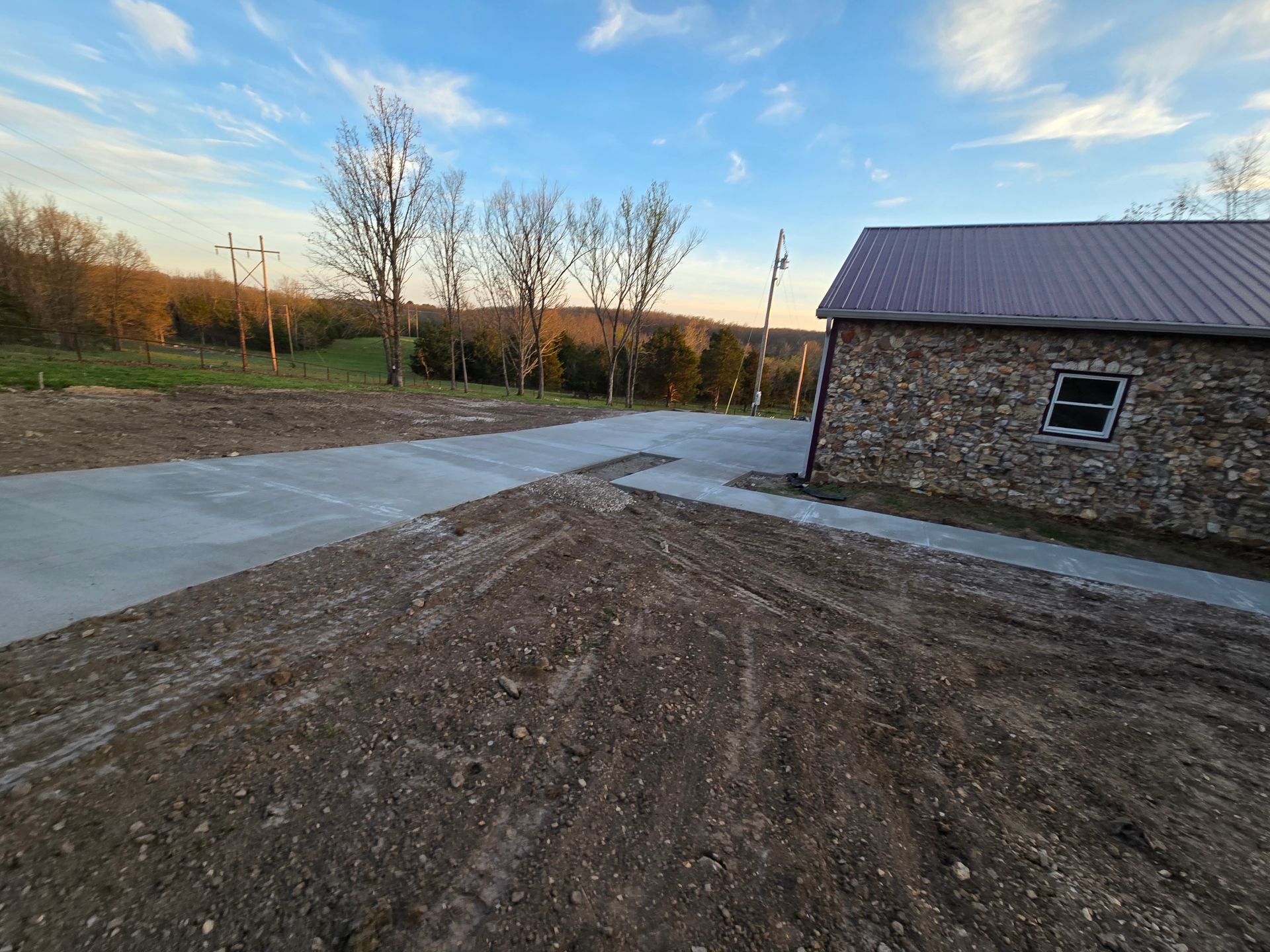 Gravel driveway beside a stone building, with trees and open land at sunset.