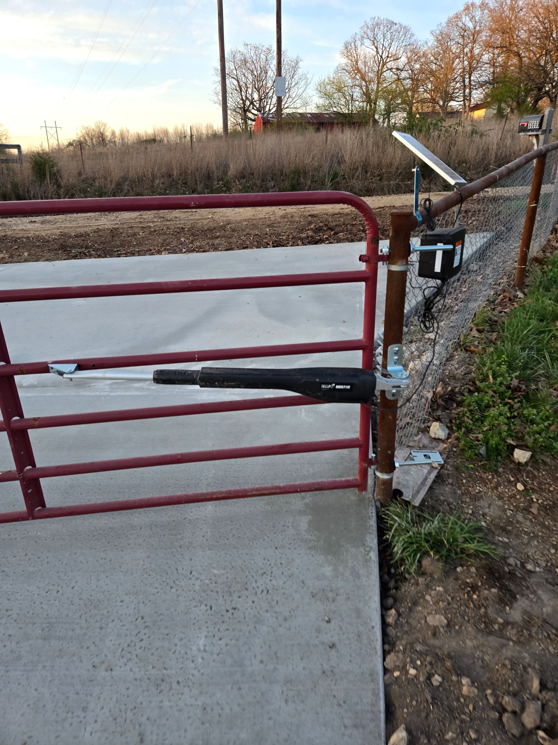 Red metal gate beside a concrete path, with a hydraulic closer and chain-link fence on the right.