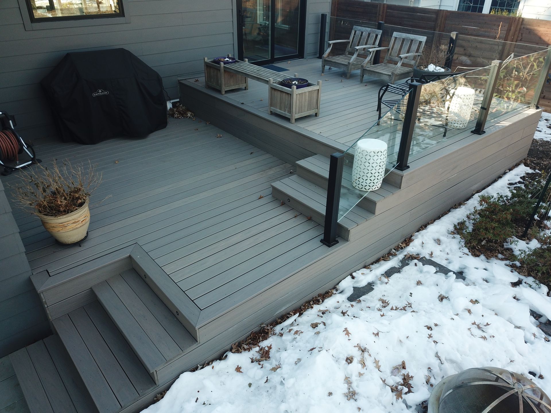 Snow-dusted backyard deck with patio furniture, grill, potted plants, and glass railing