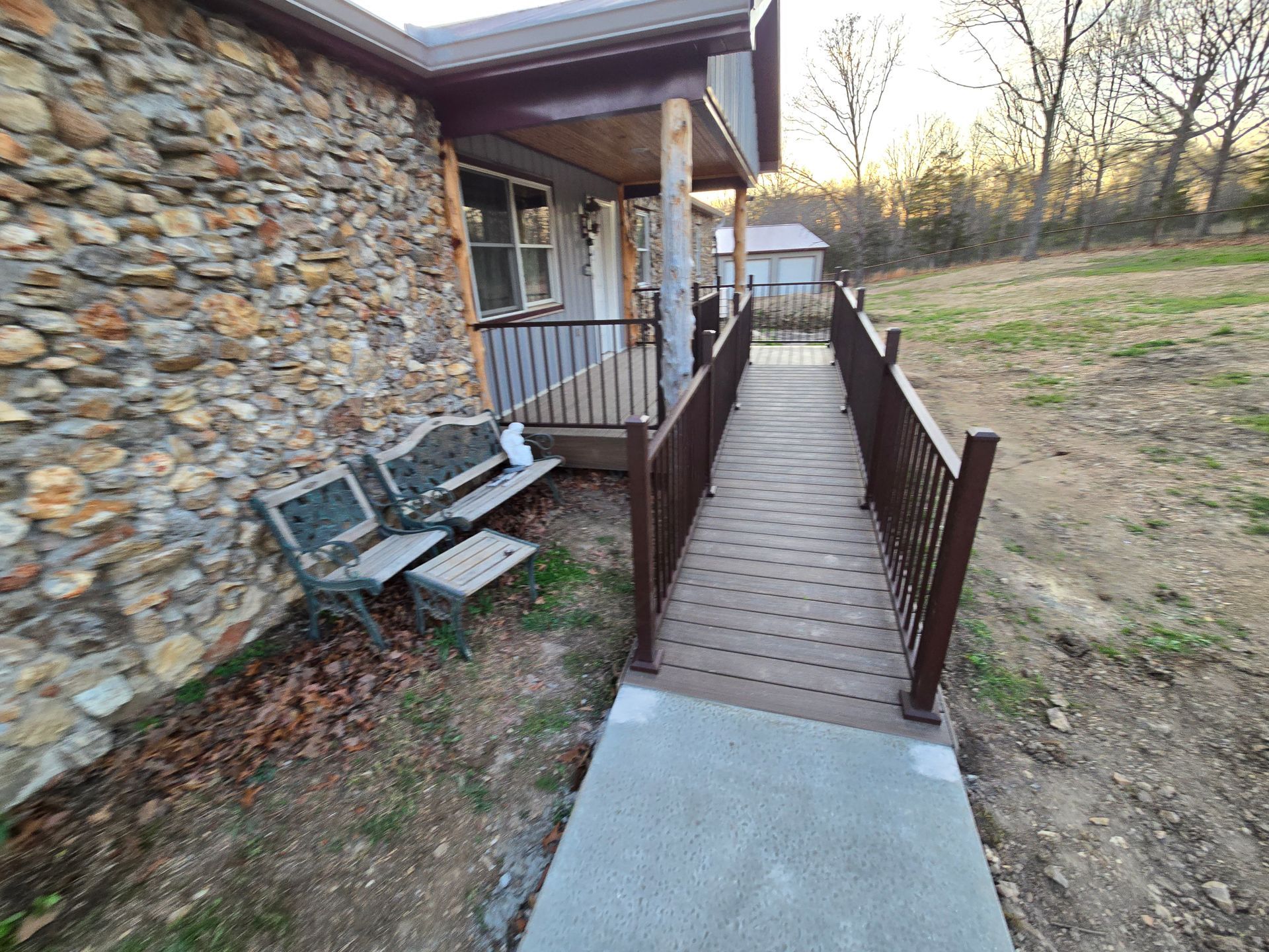 Wooden accessibility ramp beside a stone house, with benches along the wall and a grassy yard.