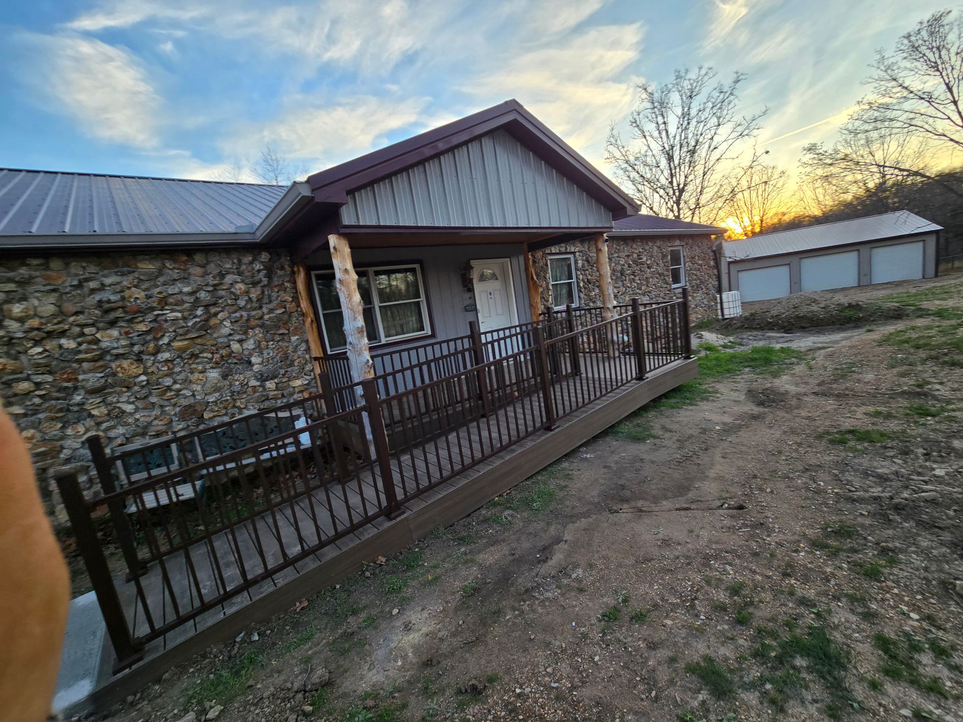 Stone house with metal roof, black fence, and dirt yard at sunset