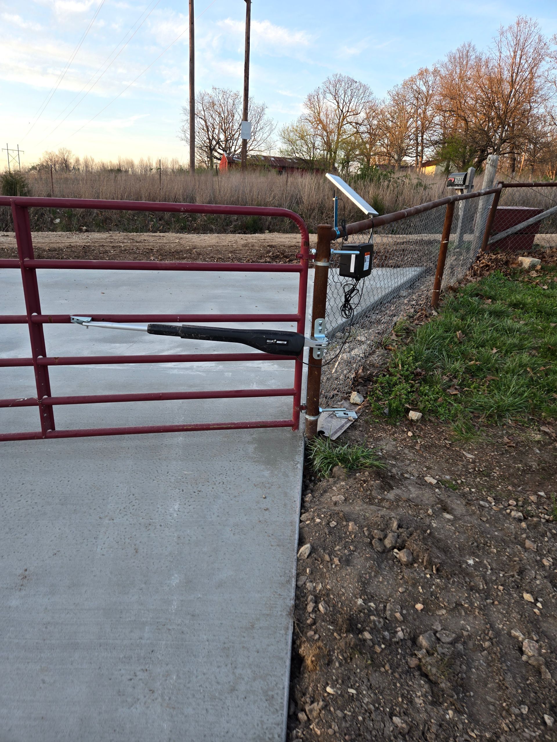 Red metal gate blocking a concrete path beside a gravel slope and chain-link fence at sunset