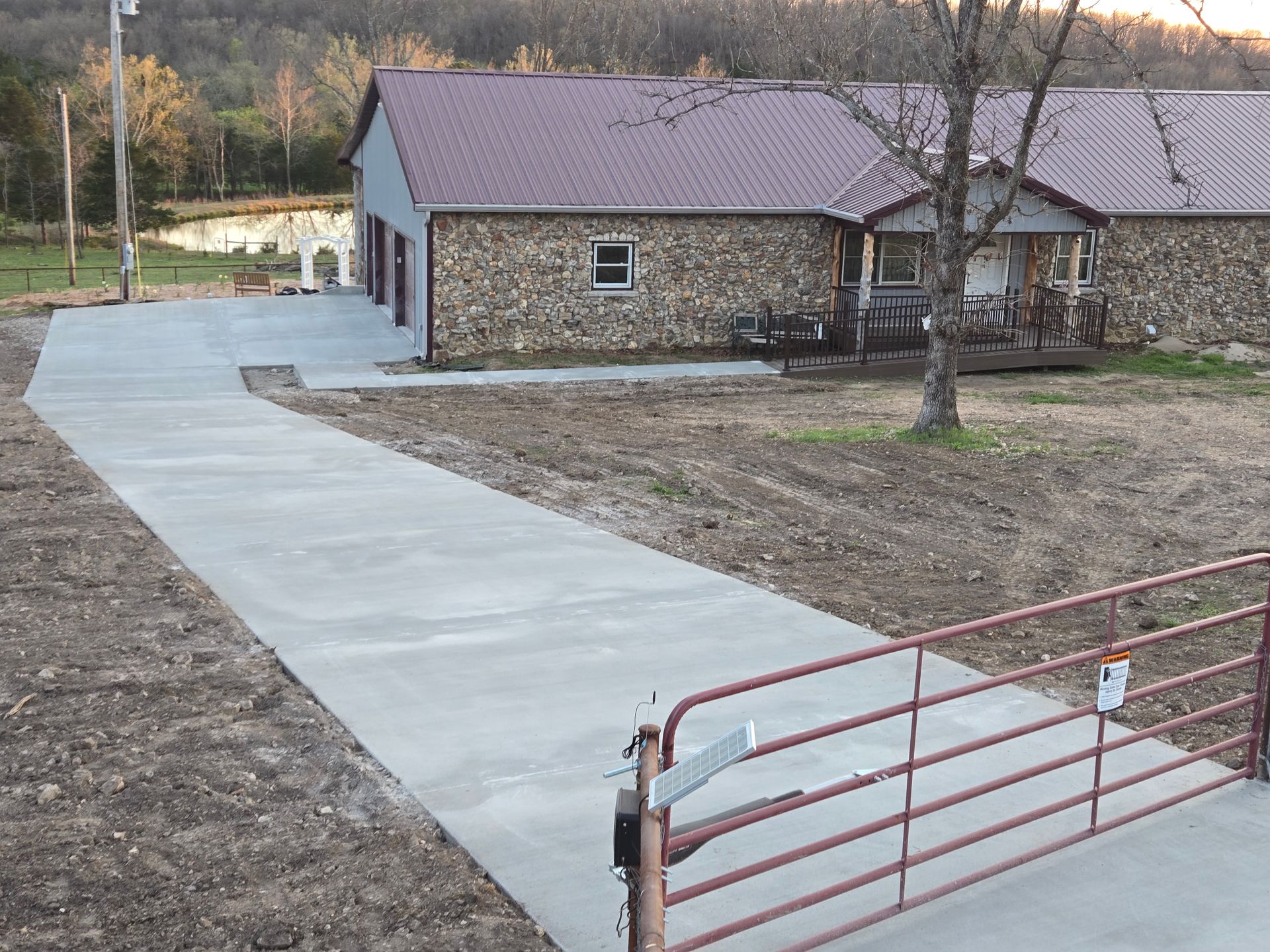 Concrete driveway leading to a stone house, with a red metal gate in the foreground.